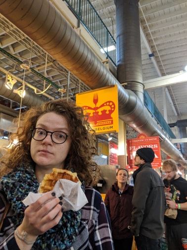 Image of Leann holding a good waffle in front of Taste of Belgium looking at the camera. 